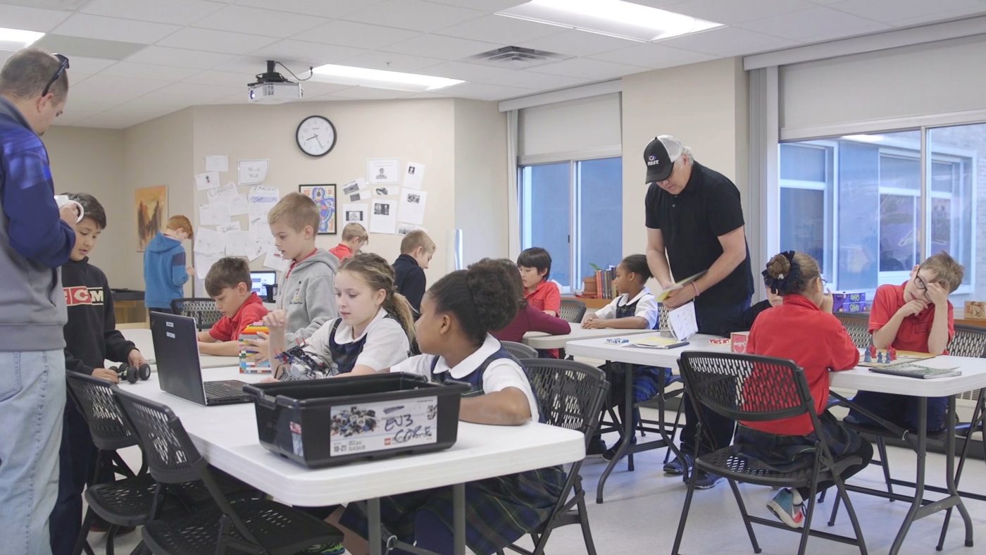 A classroom of student teams working at tables while two mentors circulate.