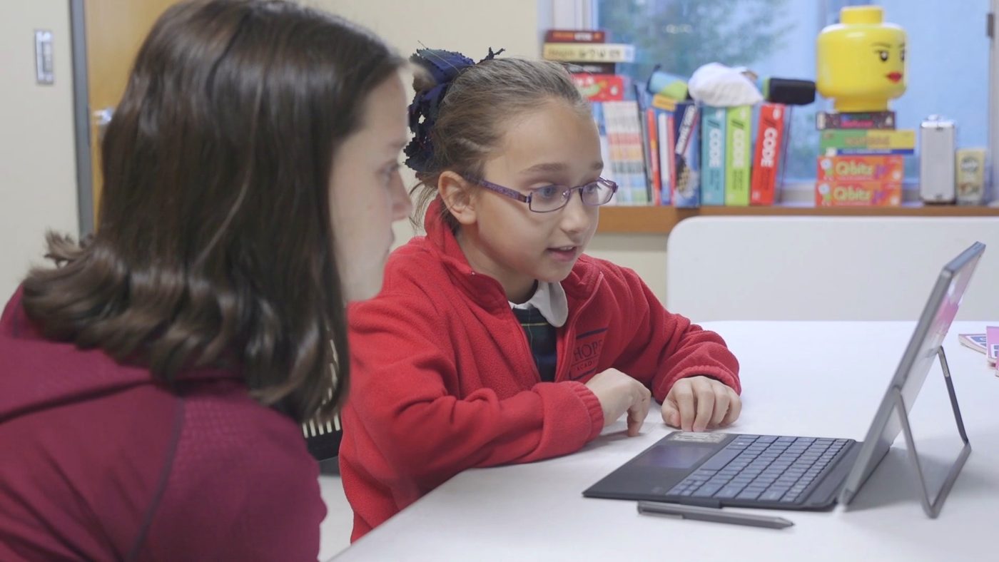 A student explains her project to a classmate at a table with a tablet.