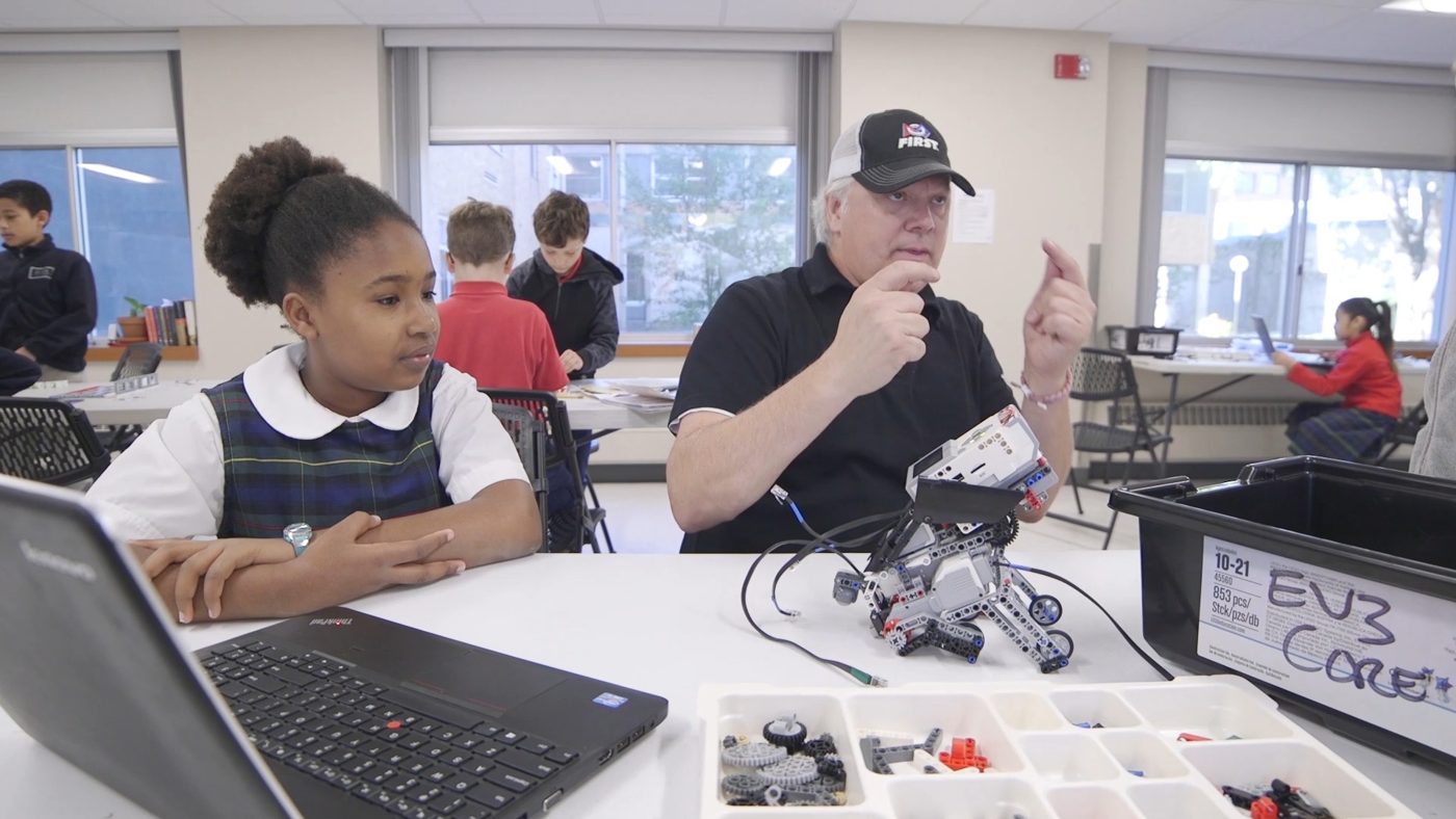 A volunteer mentor holds up a LEGO robot while a student watches from her laptop