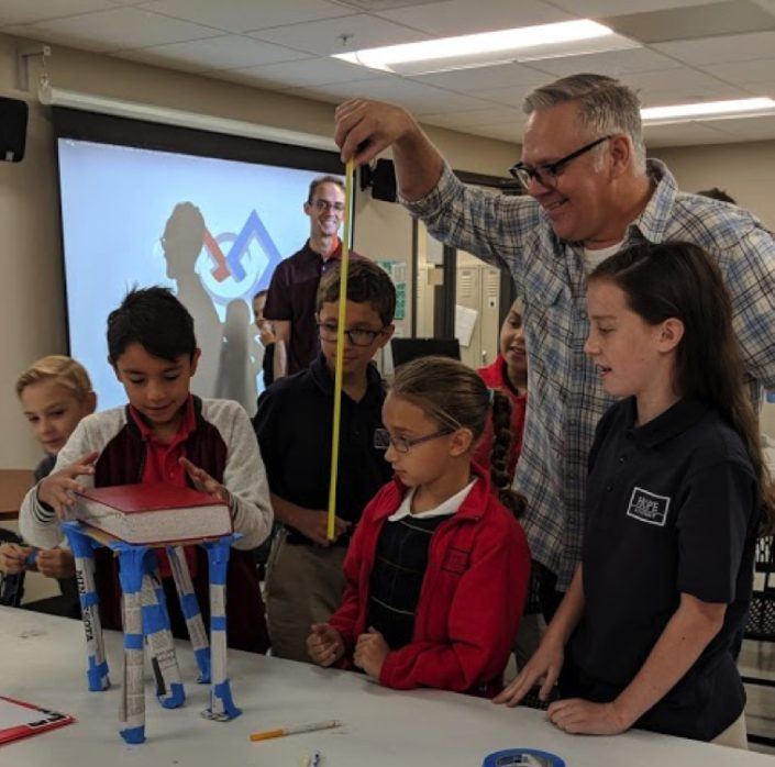 A mentor measures a student-built paper tower while classmates watch.