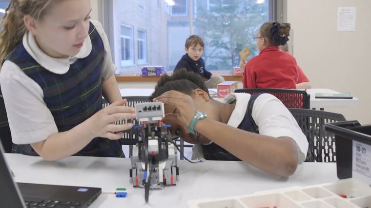 Two students adjust a LEGO robot on a classroom table.