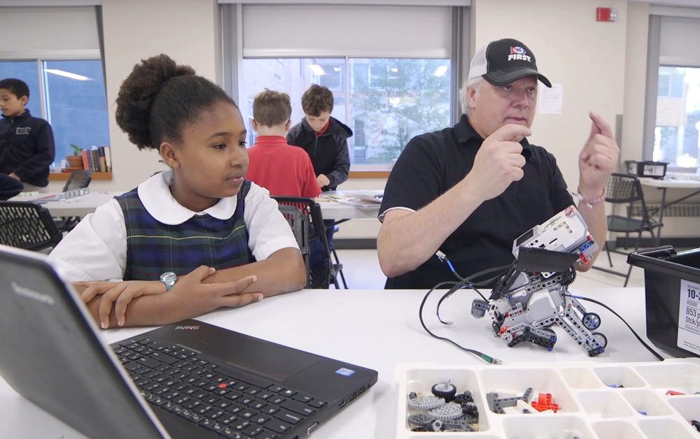A volunteer mentor in a FIRST cap talks through an EV3 robot with a student.