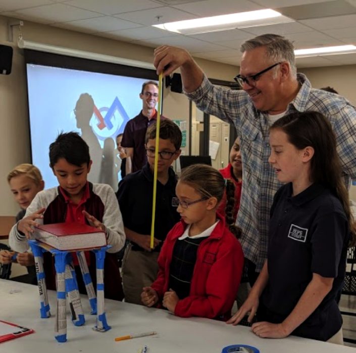 A mentor measures a tower of rolled paper tubes holding up a heavy book while students watch.