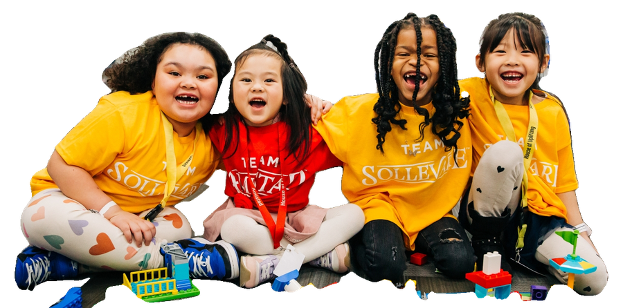 Four young students laugh together on a classroom floor, surrounded by building blocks.